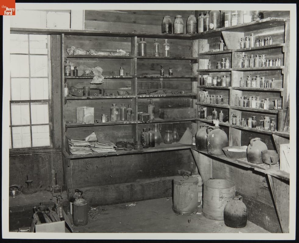 Corner of simple wooden room with narrow shelves on the walls lined with bottles, jars, and papers
