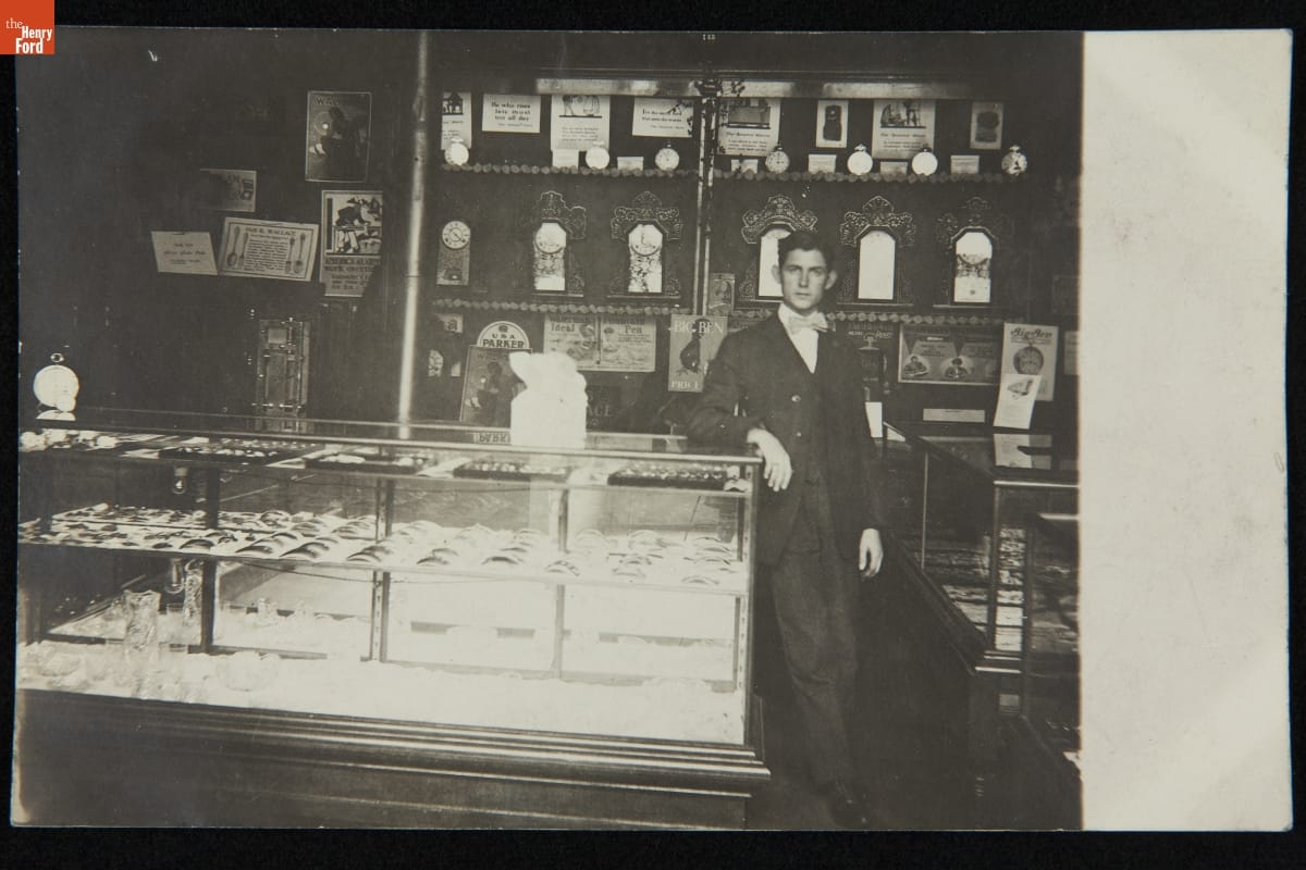 Man in suit and bowtie stands in store with arm on display case and clocks on shelves behind him