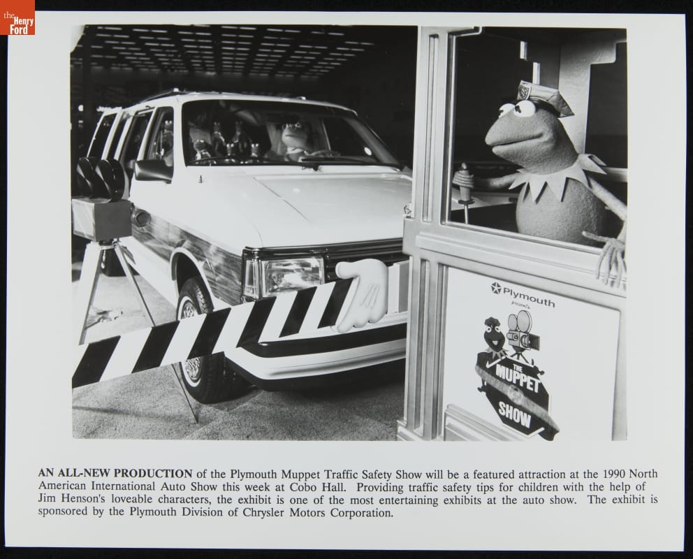 Van containing Muppet characters stopped behind barrier; Kermit the Frog in a booth in foreground; contains text