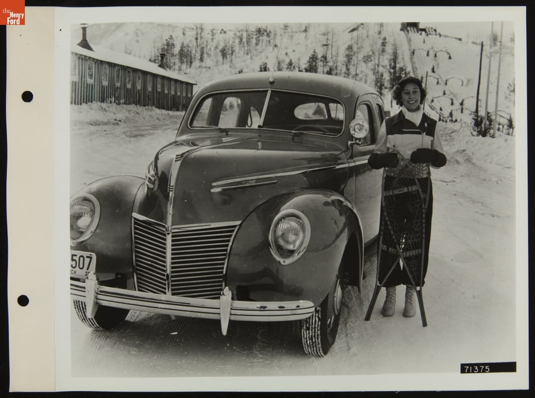 Grayling Winter Sports Queen with Mercury V-8, January 1939 Woman, holding two snowshoes, stands next to a car with a snowy hill (perhaps a ski slope) in the background and a long low wooden building to one side
