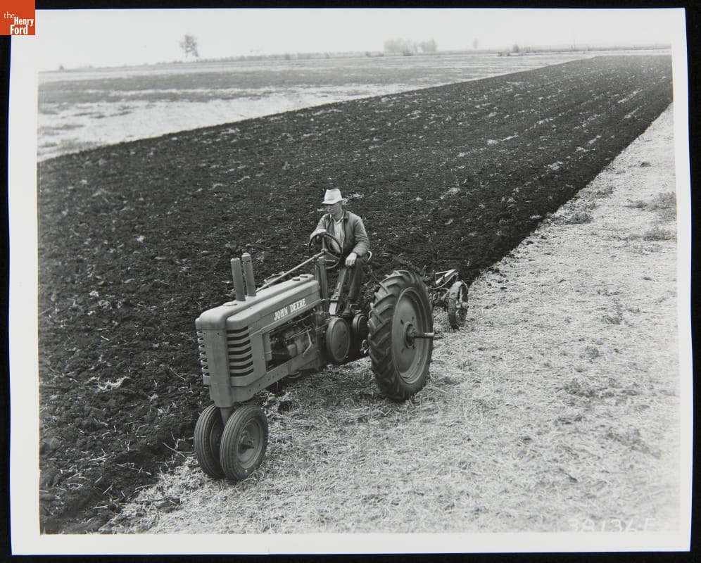 Man Using a 1939-1946 John Deere Model Black-and-white photo of man riding a tractor through a field partially dirt and partially stubble
