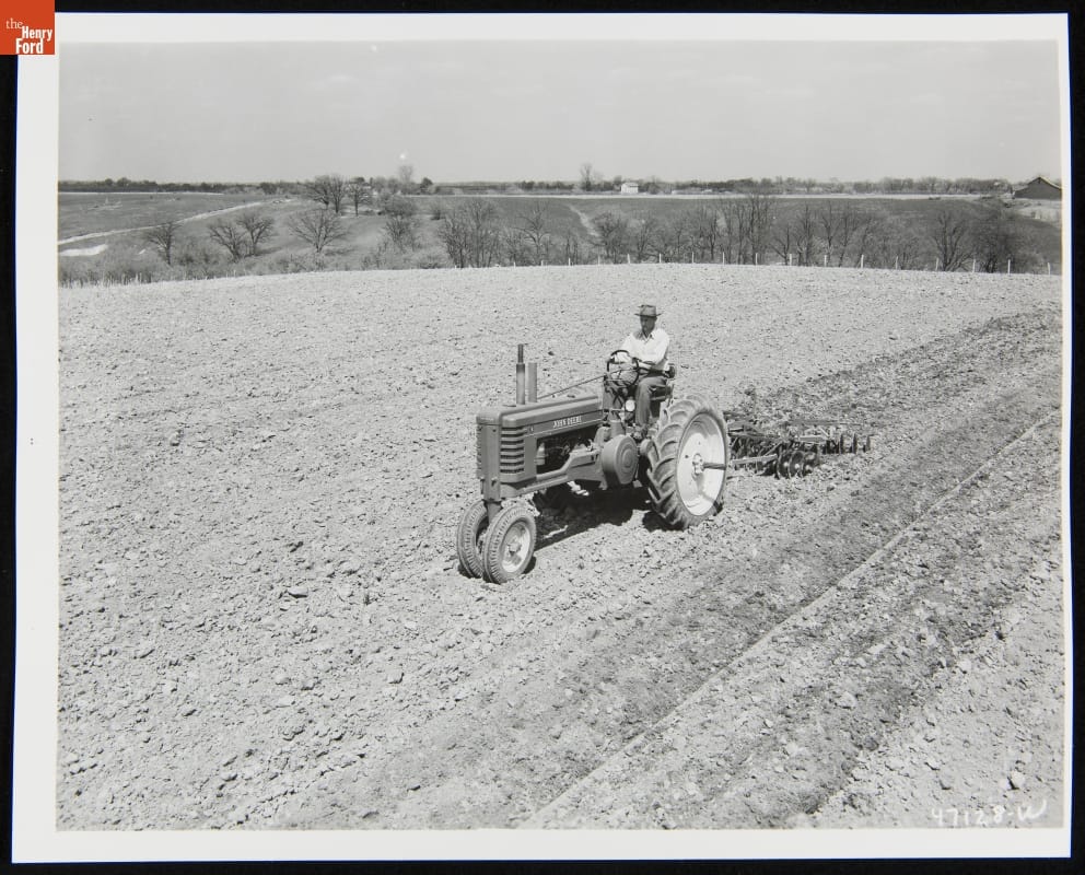Man Using a 1947-1952 John Deere Model Black-and-white photo of man riding a tractor through a dirt field