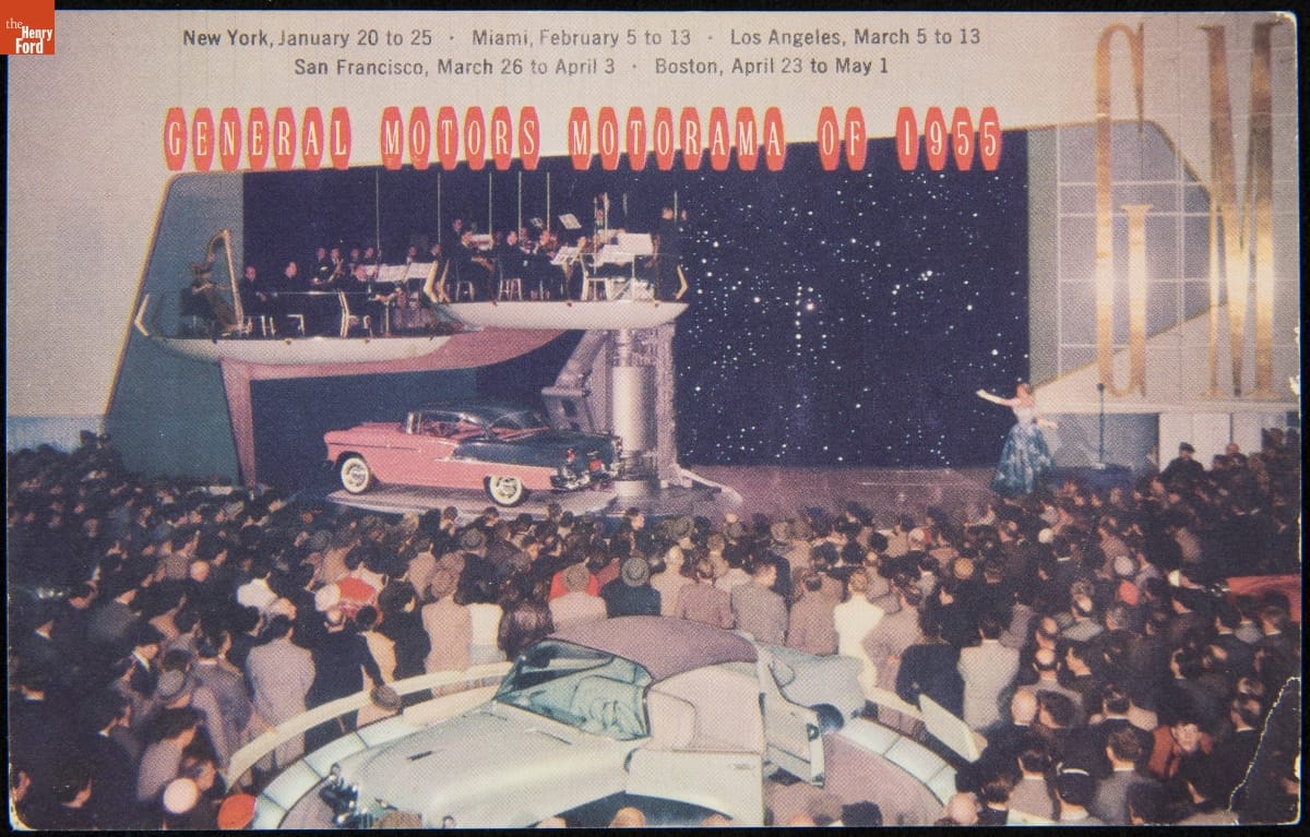 Pink and gray car on platform under another platform with musicians; a crowd of people and another car on a platform in the foreground; contains text