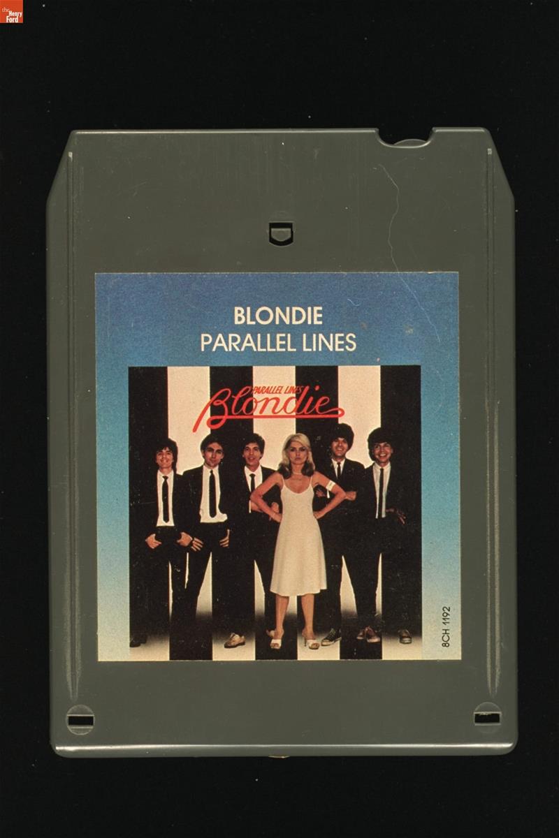 Boxy gray cartridge with square label containing text and photo of five people in front of a black-and-white striped background
