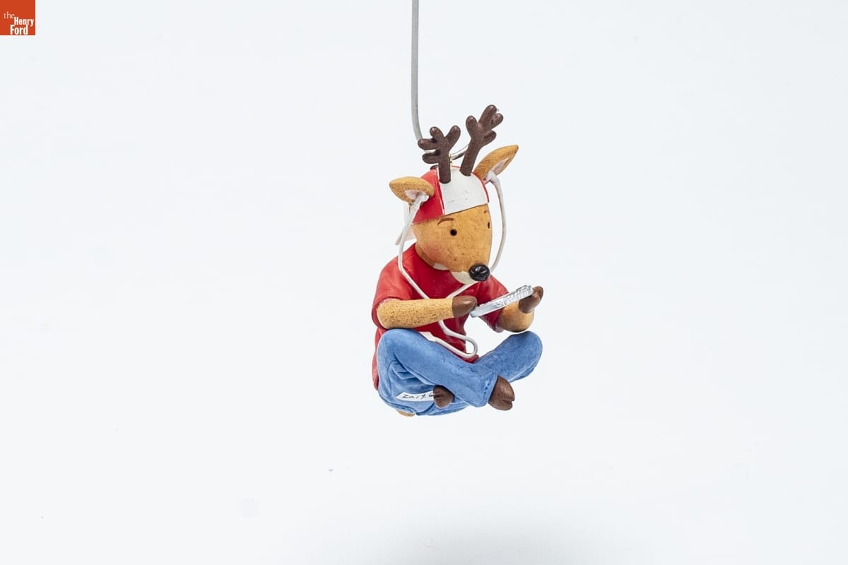Christmas ornament showing a reindeer wearing a red tshirt, blue jeans, a backwards red-and-white baseball cap, and earphones, sitting cross-legged while looking at a handheld device