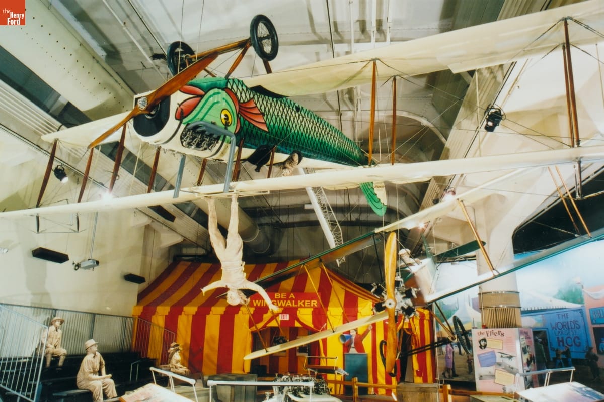 Plane with white wings and body painted like a green-scaled fish, hanging upside from ceiling in front of yellow-and-red striped tent