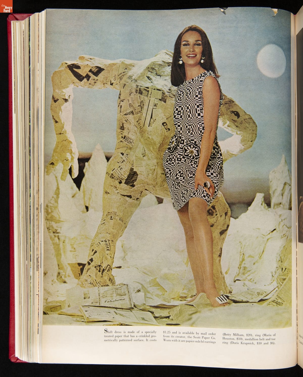 Woman in black-and-white geometric-patterned dress in front of what appears to be a papier mâché sculpture of a person in a landscape