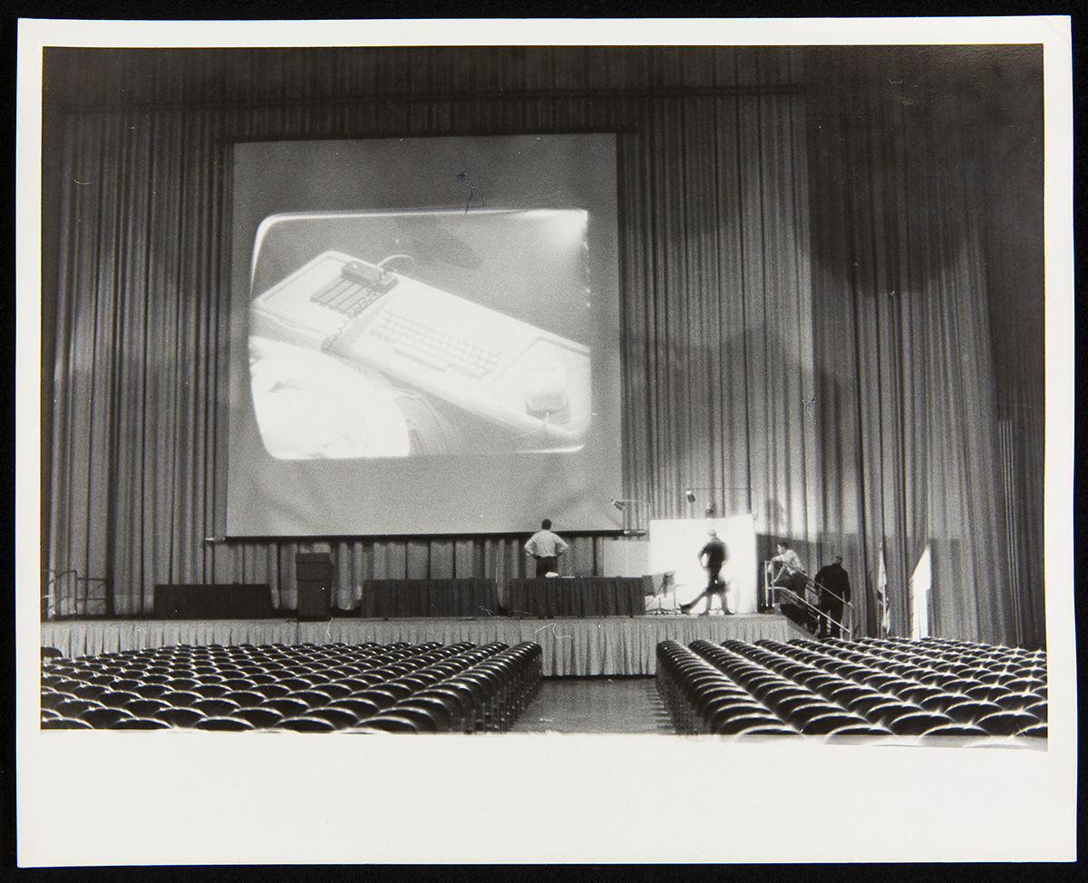 Empty auditorium with rows of seats and stage with curtains, a large screen with a piece of equipment projected on it, and several people on the stage