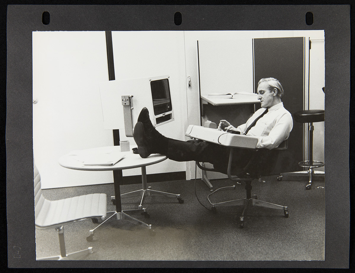 Page from three-ring binder containing photo of man sits in chair with attached console with feet up on a nearby table that also holds what appears to be a large monitor; office furniture around him