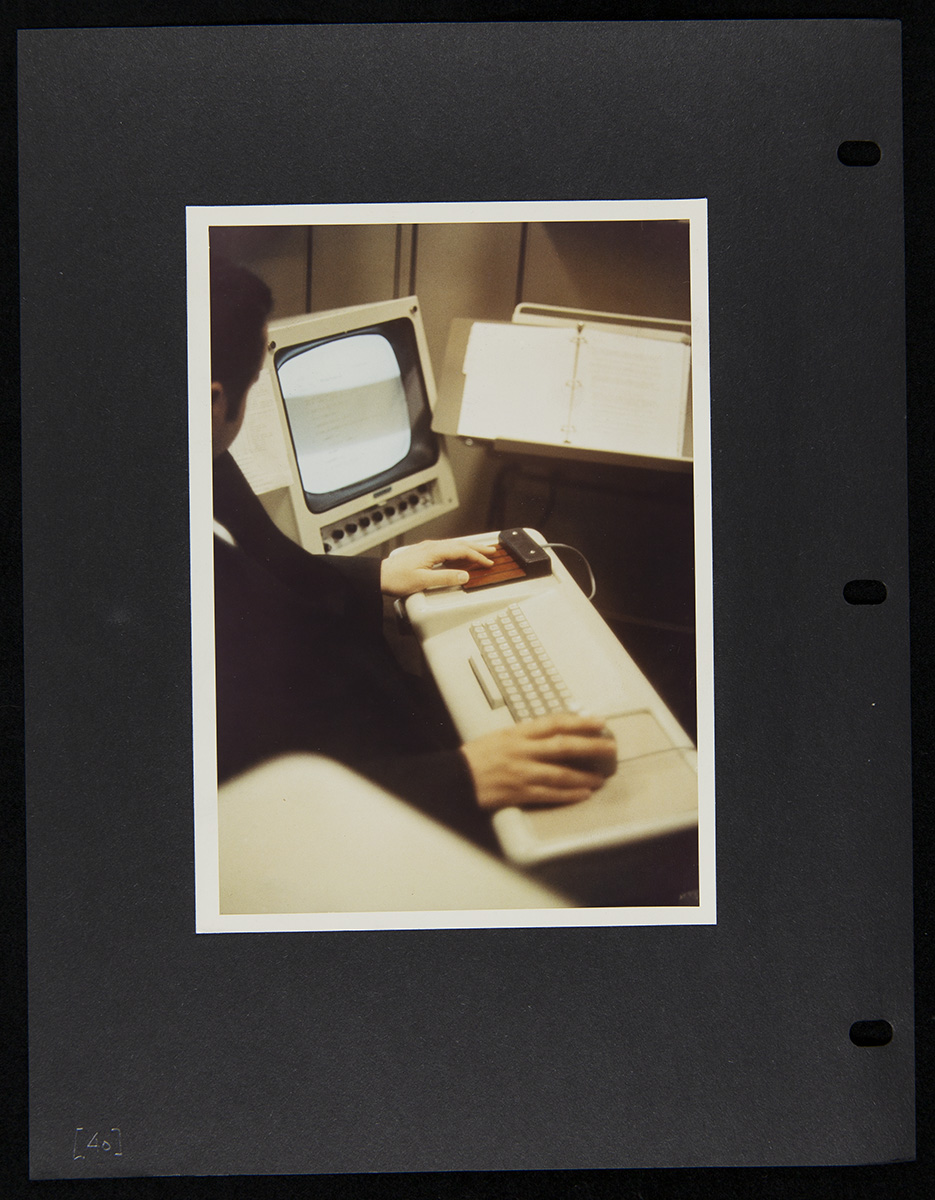 Photo pasted onto black three-ring binder sheet depicting a person in front of a keyboard with a monitor to the side and copystand in front of them