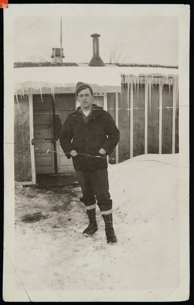 Man in coat and boots stands in snow outside simple structure covered in snow with icicles handing from eaves