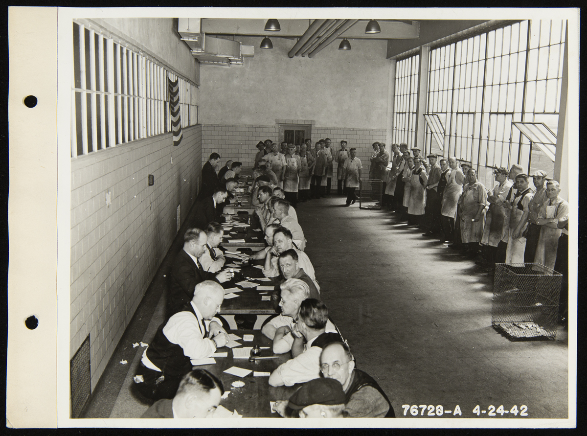 Workers Lining up at the Ford Rouge Plant for Draft Registration, April 24, 1942 Large room with small tables lined up along one side, each with people sitting on either side, and a line of people snaking around the other side of the room