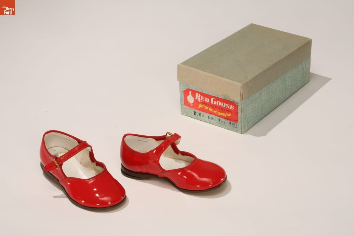 Shiny red shoes with a strap that would fall just in front of the ankle; blue shoebox next to them