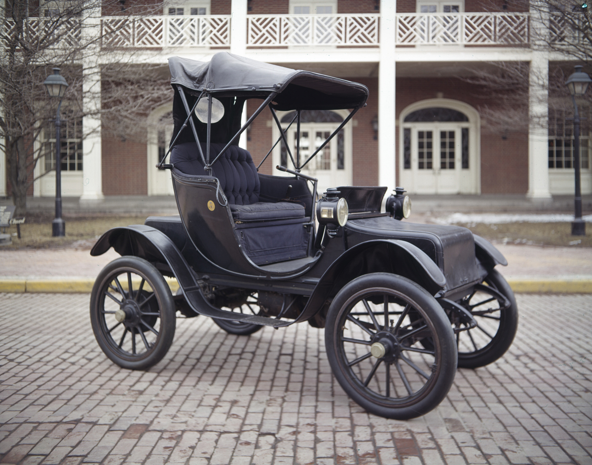Small, largely open two-seater early automobile parked in front of a red brick building with white columns