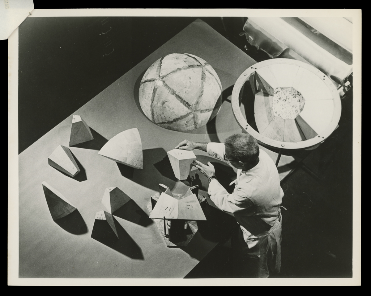 Photo from above of person in lab coat or apron working at table with hollow sphere in two pieces and various conic sections