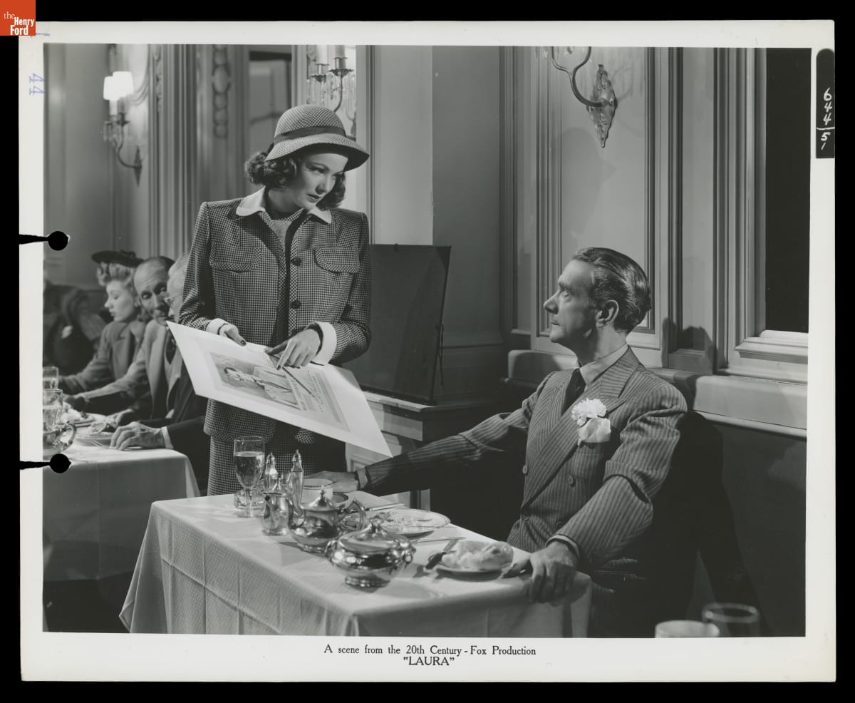 A Scene from the 20th Century-Fox Production Black-and-white photo of a woman in a suit and hat standing at a table in a restaurant showing a poster to a man seated at a table