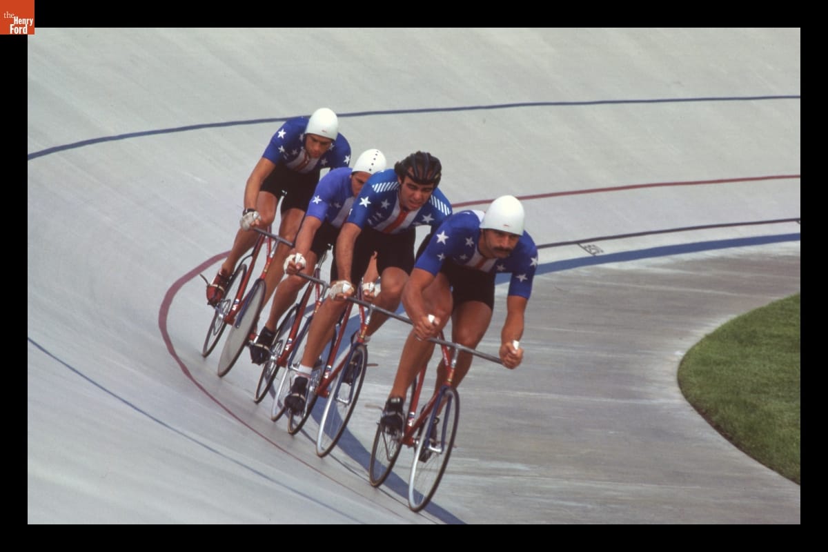 Four cyclists in American flag inspired outfits riding in single file on a track.