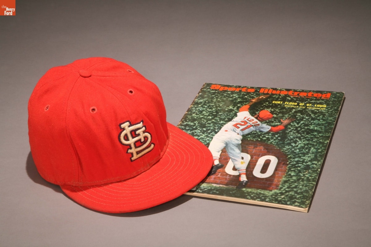Red baseball cap with logo on front and Sports Illustrated magazine with image of baseball player jumping for ball on cover