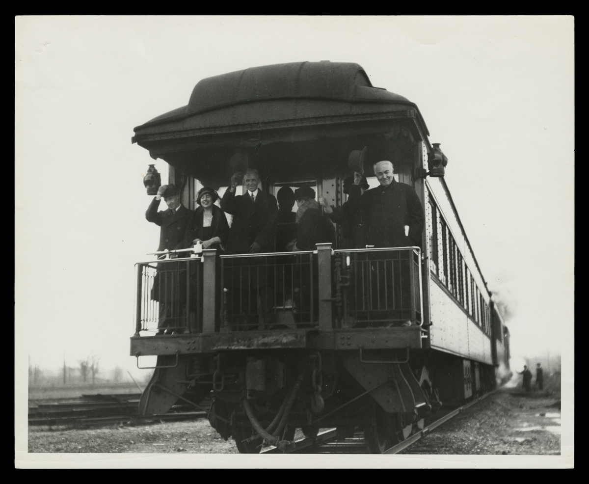 Edsel and Eleanor Ford, Henry and Clara Ford, and Mina and Thomas Edison on the Private Railroad Car Black-and-white photo of six people on the back platform of a rail car, most of them smiling and waving