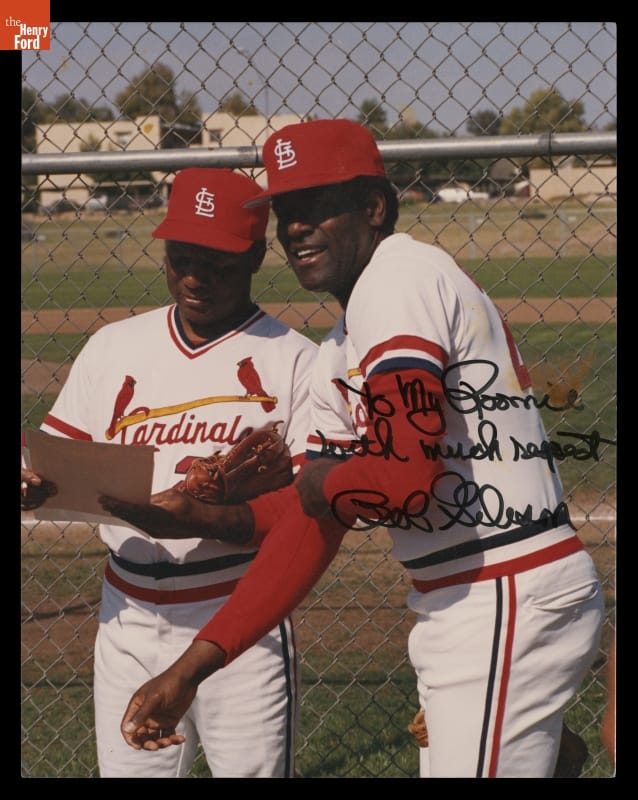 Two dark-skinned men in red and white baseball uniforms stand in front of a baseball field