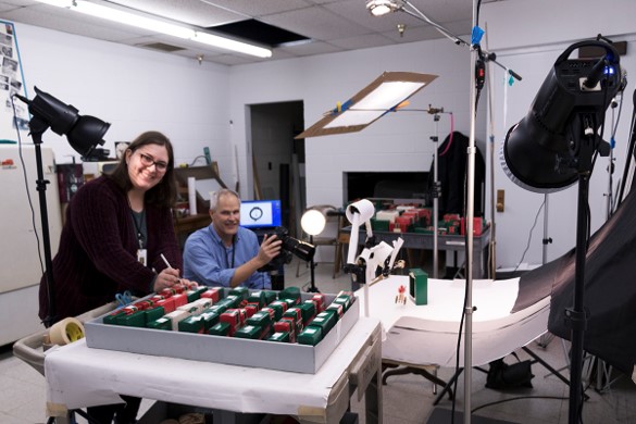 Contractors Todd Seroka and Natalie Diehl photographing Hallmark ornaments Man points camera at photo set and woman works at cart filled with small green and red boxes