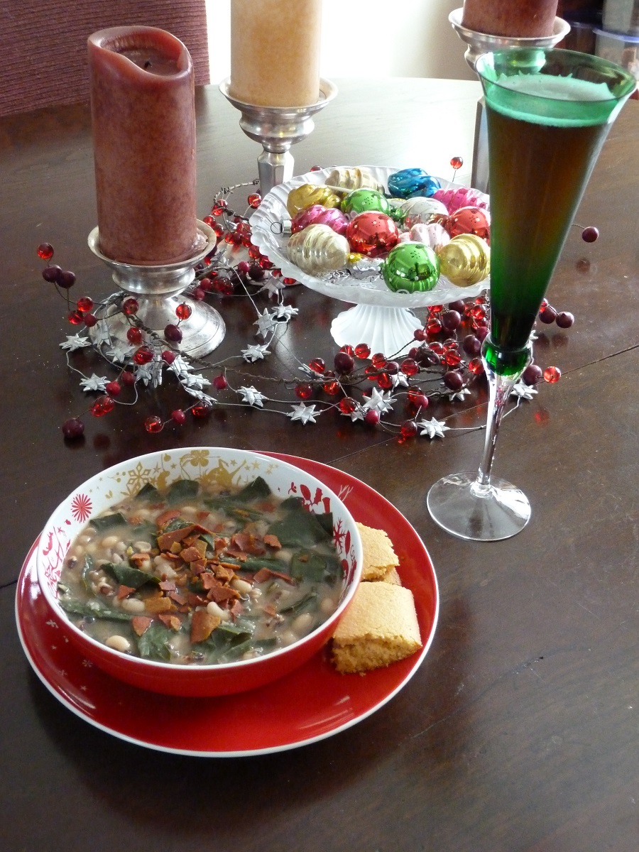 Red dishware containing a soup with greens and veggie bacon, with cornbread on the side, a flute filled with liquid, and a holiday centerpiece with candles, ornaments, and garland